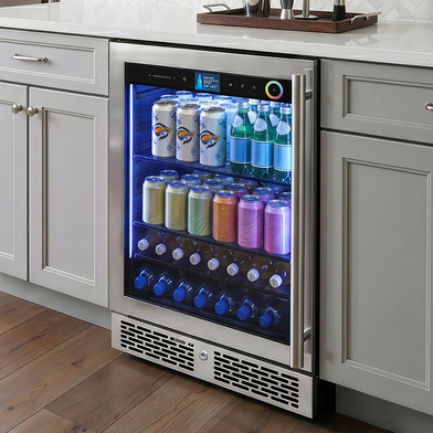 A stainless steel undercounter beverage refrigerator installed in a kitchen cabinet, stocked with assorted canned and bottled drinks. The refrigerator features a digital control panel, blue LED lighting inside, and a bottom vent grille, set against light gray cabinets and a white countertop.